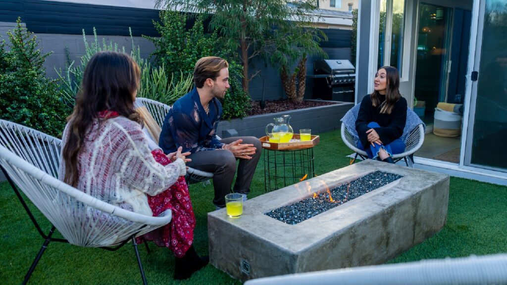 a photo of three people sitting around a firepit in a small backyard with turf