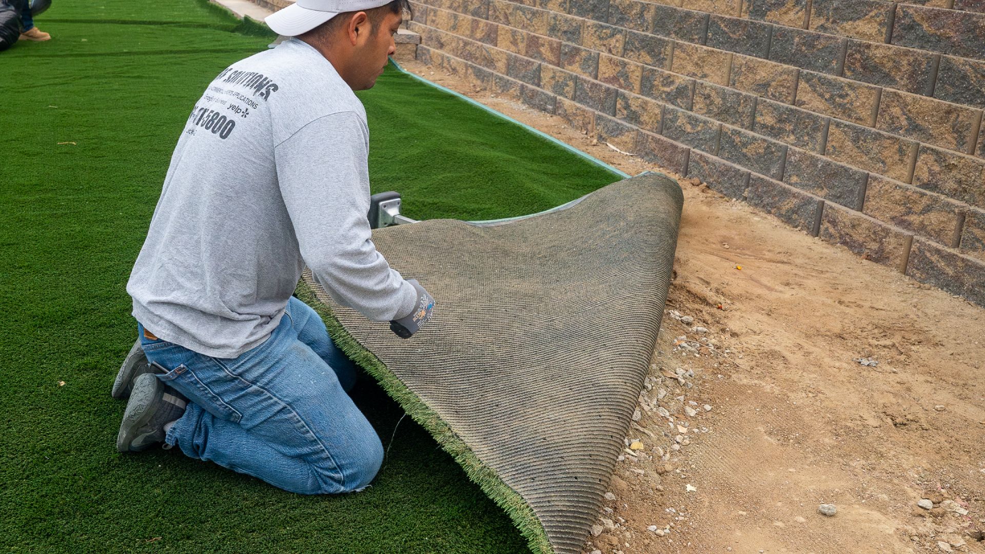 a photo of a turf installation expert laying down turf in a backyard next to a stone wall